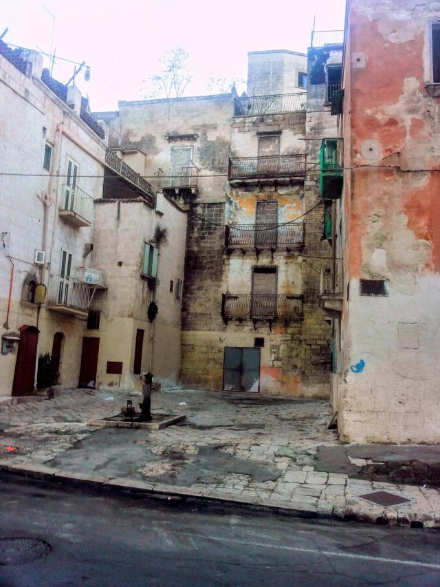 Un largo della Città Vecchia di Taranto con palazzi dalle facciate scrostate, balconi in ferro e una fontanina